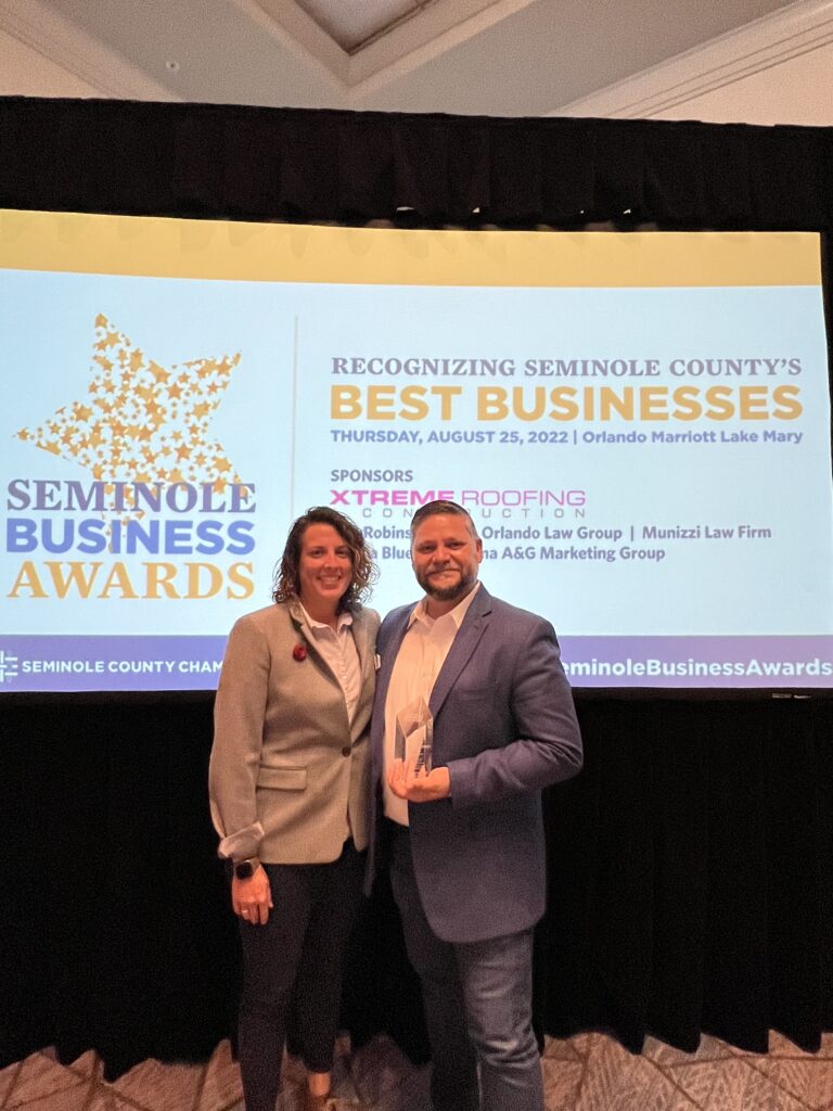 Co-owners of Solcium Solar, Michelle Solano and Gustavo Panosso, accept the award for 2022 best large business in seminole county in front of a Seminole Business Awards screen