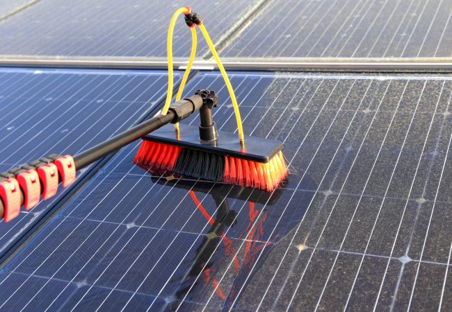 A brush with water glides across an array of solar panels as it gently cleans off the debris and pollen that is visible on some of the panels.